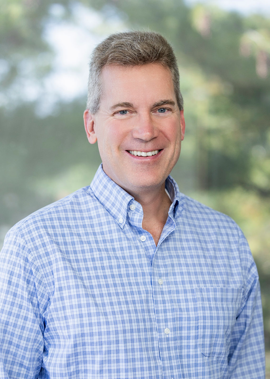 Man in a blue checkered shirt with short grey hair smiling against a green background