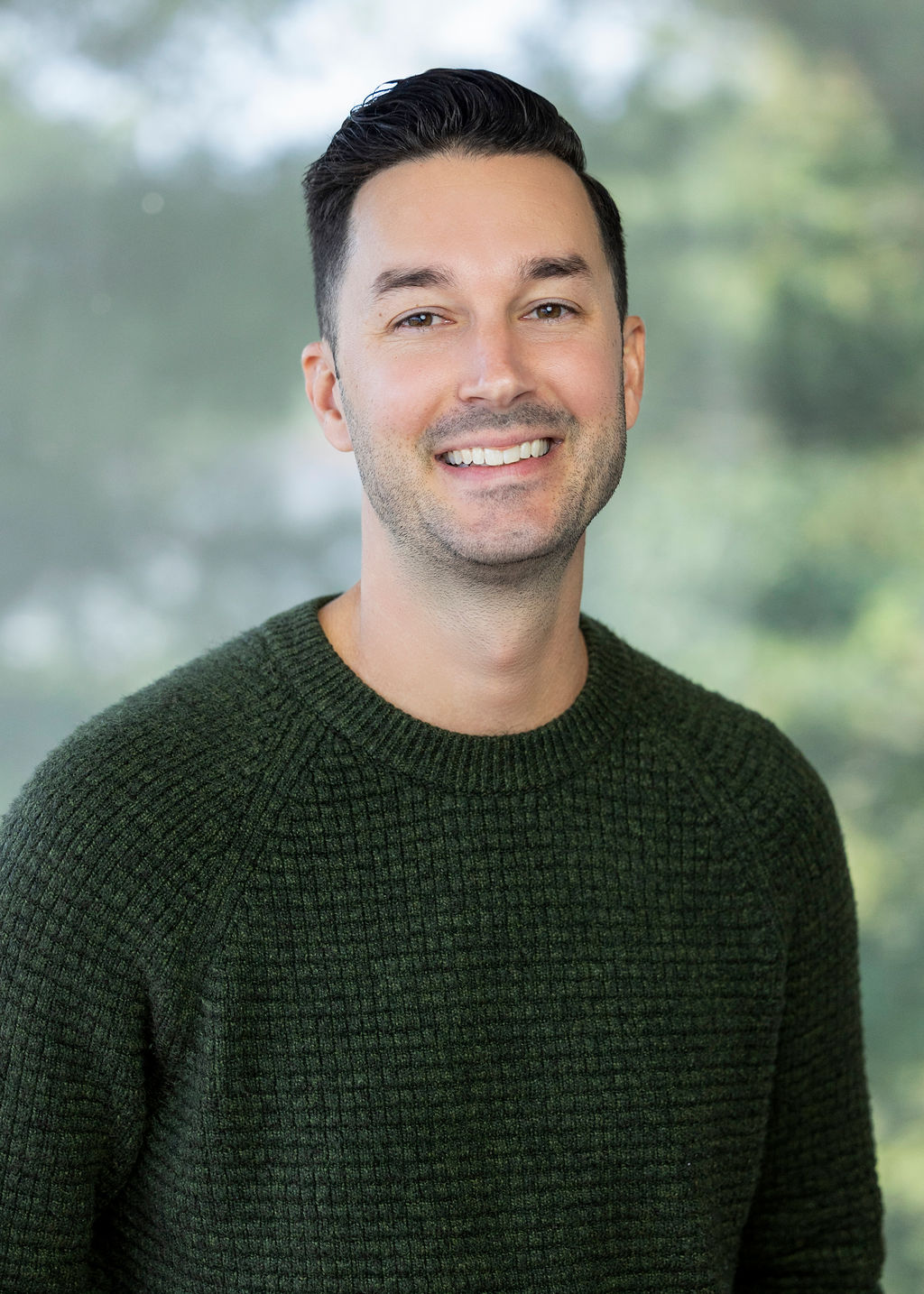 Man wearing a dark green sweater with dark brown hair smiling agains a green background