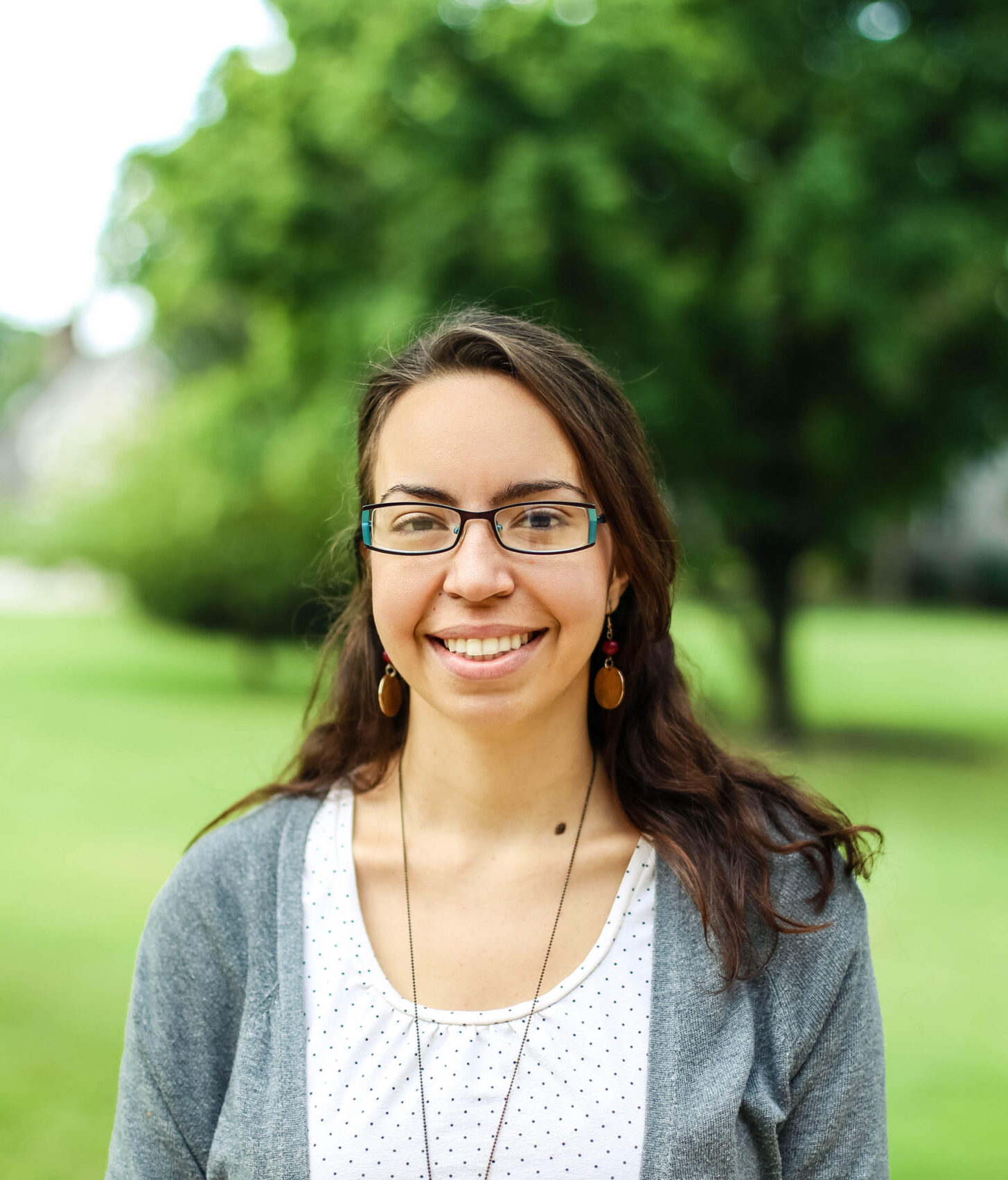 Woman in glasses with a grey cardigan and white shirt smiling against a blurred nature background