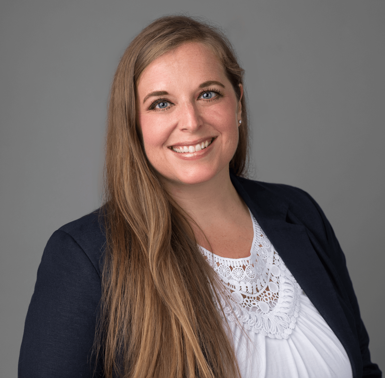 Woman in a black blazer and white blouse with long blonde hair against a grey background